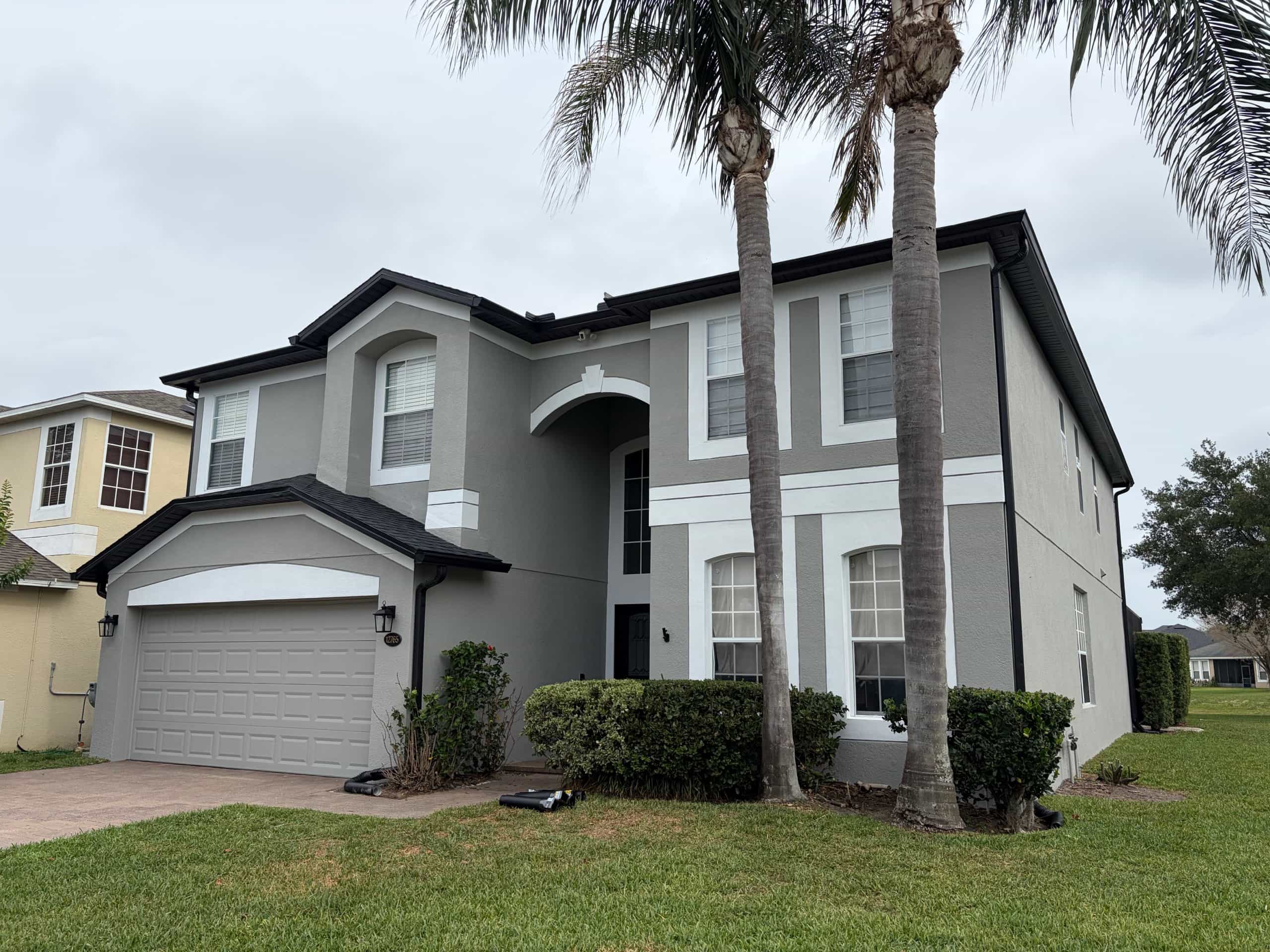 Two-story home exterior repainted sage green with white trim banding and black fascia, framed by twin palm trees