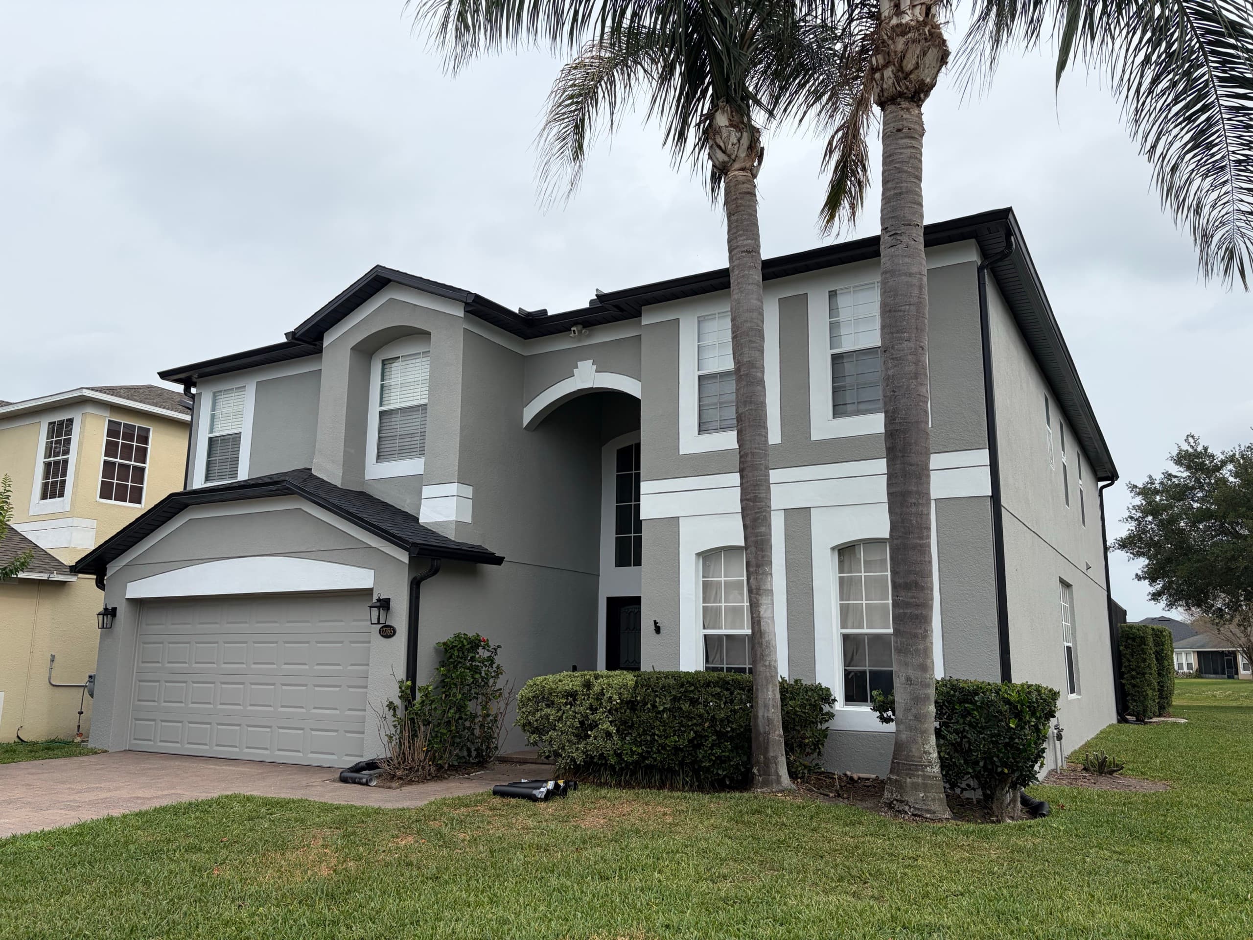 Two-story home exterior repainted sage green with white trim banding and black fascia, framed by twin palm trees